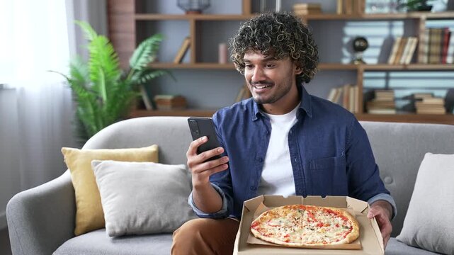 Satisfied young man holds an open box with pizza sitting on sofa in living room and writes good review on phone. A smiling handsome man is happy about fast delivery of food. Ordering food to take home