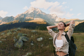 Naklejka premium Young woman with a backpack stands in a mountain landscape, smiling with arms raised as sunlit peaks rise behind her and a wide blue sky. Under clear weather and fresh air.
