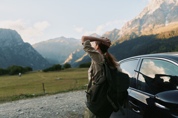 Fototapeta premium Young woman with backpack stretches her arms beside a parked car in a rugged mountain valley, exuding curiosity and freedom with a sunlit dramatic landscape behind.