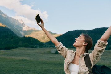 Naklejka premium Joyful woman outdoors, taking a selfie with a phone, in a sunny meadow with a mountain backdrop, relaxed posture, natural colors, fresh breeze.