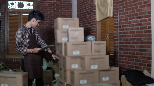 Worker checking inventory beside stacked boxes with tablet and clipboard brick wall storeroom, apron, labeled cardboard packages,