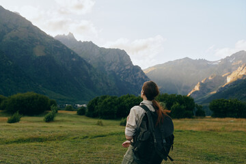 Fototapeta premium Young woman with backpack stands in a sunlit valley, gazing toward distant mountains, capturing a moment of exploration and calm amid nature.