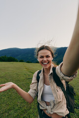 One person young woman with backpack takes a sunny meadow selfie with mountains behind, smiling...