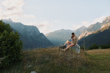 Fototapeta premium Woman enjoying outdoor lifestyle in a mountain meadow, sitting on a rock and gazing at distant peaks, casual clothes, peaceful mood, warm sunlight, natural landscape and solitude inspires calm.