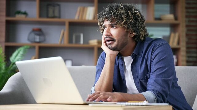 Bored young adult man sitting in home office, looking at laptop with a weary expression. Businessman feeling unenthusiastic. Frustrated male entrepreneur procrastinates. A lazy student gets distracted