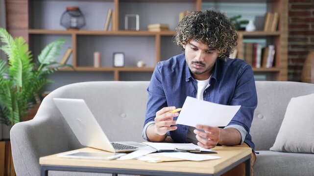 Confident young adult man dealing with household finances while sitting on sofa in home office. Busy male calculates busy with paper work, calculates budget, takes notes, fills out form of documents