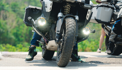 A close-up of a motorcycle's front wheel and headlights with the rider"s feet on the ground, parked on an asphalt road surrounded by greenery. © PIPAT