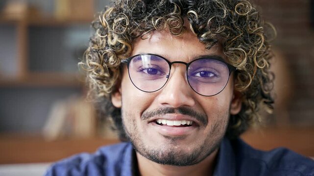 Portrait of happy handsome man sitting on sofa in living room at home. Head shot of confident smiling male in glasses. Positive friendly man with curly hair poses looking at camera indoors. Close up