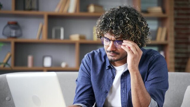 Young man suffering from eye fatigue and headache while working on a laptop sitting at home office. Overtired entrepreneur takes off his glasses and massages bridge of his nose with his hand. Close up