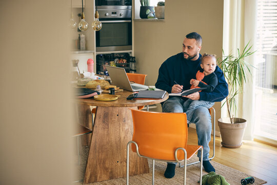 Focused man writing notes through laptop while sitting with daughter at table in home office