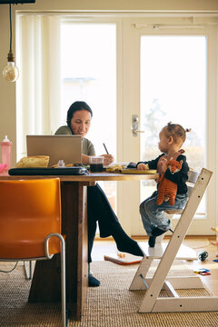 Full length of girl playing with toys while sitting beside mother writing notes while talking on work call at home