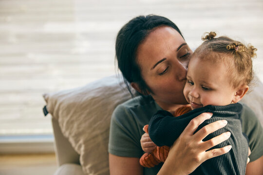 Affectionate mother kissing daughter while spending leisure time with her at home