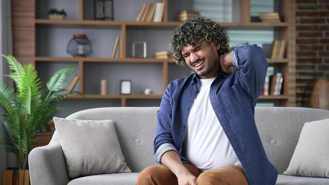 Tired young adult man suffering from neck pain sitting on sofa in living room at home. Upset male feels discomfort, massages rubs sore muscles, stretches himself. Concept of sore muscles, bad posture