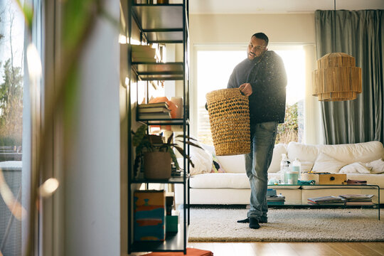 Full length of man holding laundry basket while talking on smart phone doing house chores