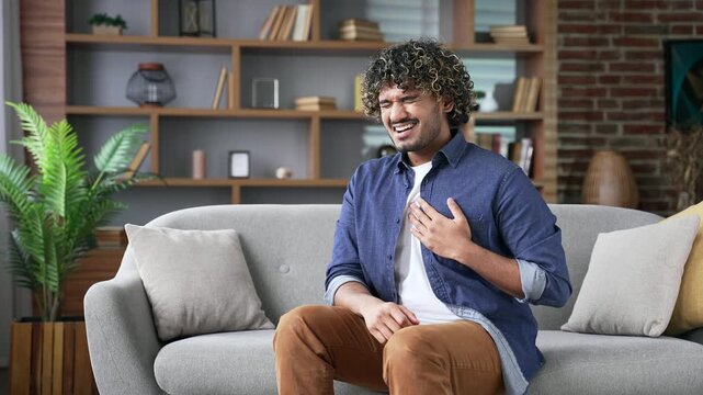 Sick adult man suffering from chest tension, heart attack sitting on sofa in living room at home. Upset handsome male holding his chest with his hands, breathing deeply, needs urgent medical attention