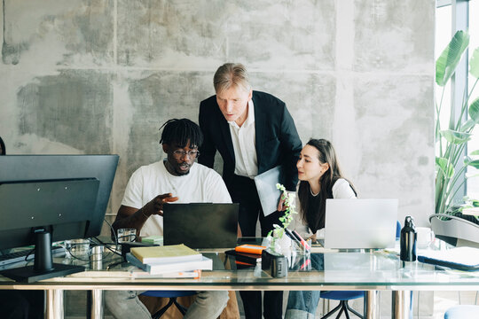 Male investor discussing business ideas with employees sitting at desk in startup workspace