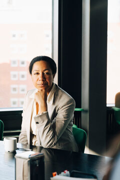 Portrait of confident female investor sitting with hand on chin at table in office meeting room