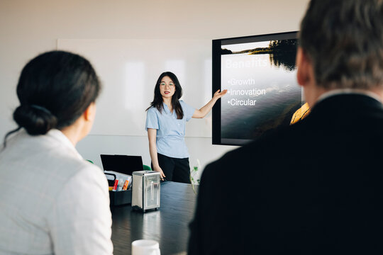 Young female employee giving presentation to investors sitting in meeting room at office
