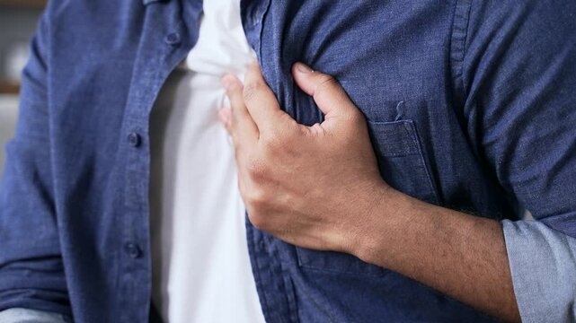 Close up. Male hands holding massaging chest. Young adult man in white shirt has heart attack while sitting on sofa in living room at home. Sick man is breathing deeply. He needs urgent medical help