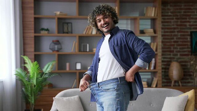 Happy handsome man feels joy after losing weight. Smiling satisfied young adult male tries on his big pants jeans, which he wore before, standing in living room at home looking joyfully at the camera