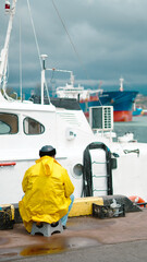 Fototapeta premium Vertical shot of a person in a bright yellow raincoat sitting on a stool at a pier, looking toward ships in a harbor.