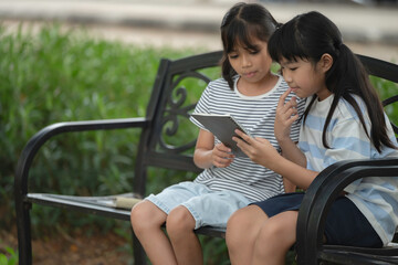 Obraz premium Happy Asian student girls reading book together on stairs at university. Back to school concept with kids sitting outdoors, smiling, studying, and learning with backpacks and headphones in summer.