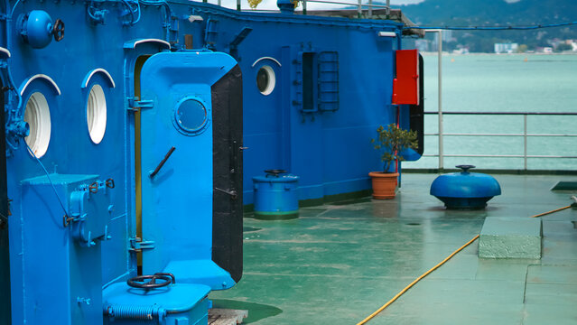Blue ship deck with an open metal door, portholes, and green flooring against a sea backdrop