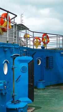 Blue ship deck with lifebuoys, an open heavy door, and portholes under a cloudy sky.