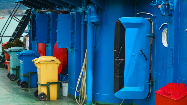 a blue ship deck featuring an open heavy door, colorful waste bins, and industrial plumbing