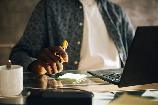 Midsection of businessman with adhesive notes and pen next to laptop on glass desk at office