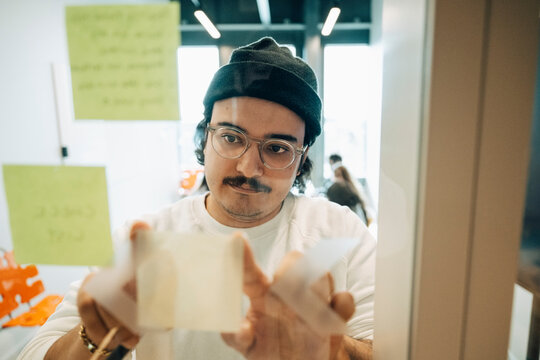 Male employee sticking adhesive note on glass wall inside meeting room