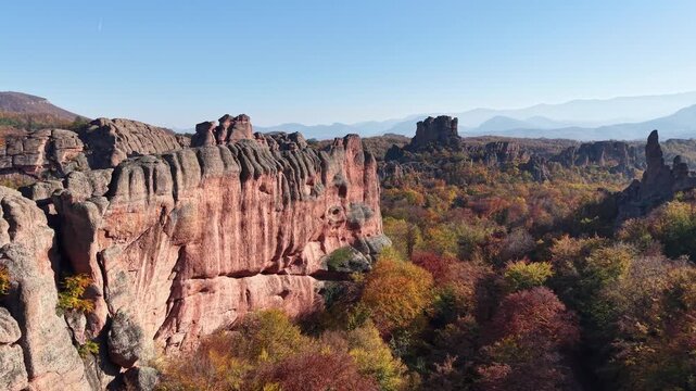 Medium-wide aerial pass along a natural red stone cliff above colorful autumn woodland and an open rocky landscape.