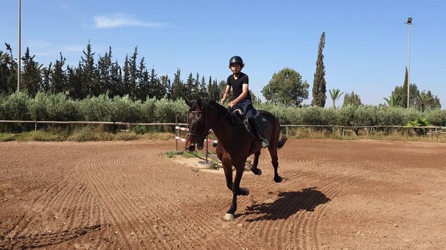 ​A dynamic, ground-level medium shot of a young equestrian rider, a teenager, cantering a powerful dark bay horse through a turn in a dirt training arena. 