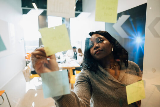 Businesswoman writing ideas on sticky notes during brainstorm session with team at modern startup office
