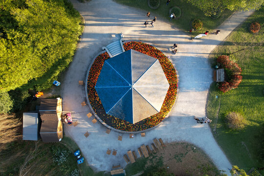 Aerial view of a circular pavilion with a blue and white roof surrounded by vibrant flower beds and people walking on gravel paths in a park Morges, Vaud, Switzerland.