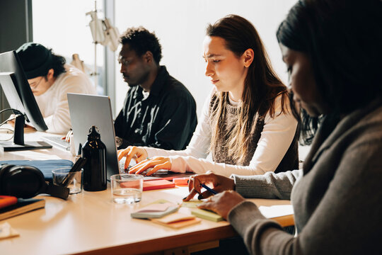 Young businesswoman working on laptop while sitting at desk with coworkers in creative workspace