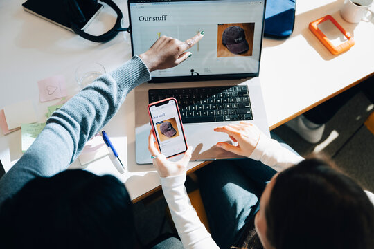 High angle view of businesswomen comparing websites through phone and laptop while sitting at desk in office