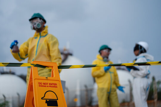 Industrial safety team in hazmat suits and respirators securing restricted chemical zone with warning sign and caution tape during hazardous materials inspection at industrial facility