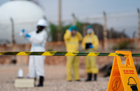 Industrial safety team in hazmat suits and respirators securing restricted chemical zone with warning sign and caution tape during hazardous materials inspection at industrial facility