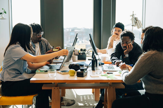 Diverse team of programmers working on laptops and computers while sitting around cluttered desk in office