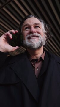 mature bearded man engages in mobile phone call while walking near modern building. Male entrepreneur focused and attentive conversating with smartphone device, enjoying communication technology 
