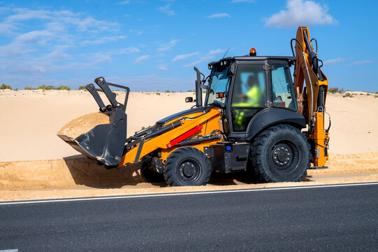 Excavator moves sand along a coastal road in Parque Natural de Corralejo, Fuerteventura, showing contrast between infrastructure work and natural dune landscape under blue sky.