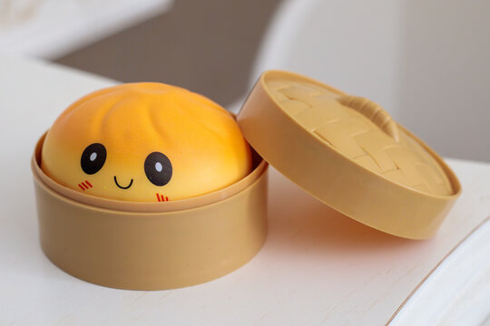 yellow kawaii squishy dumpling toy with smiley face sitting inside open plastic steamer box with lid on white table surface