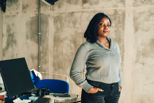 Portrait of confident businesswoman posing with hands in pockets near cluttered desk at office