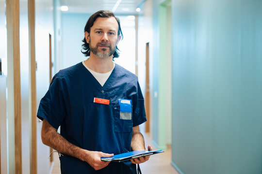 Portrait of male physician holding file folder while standing in hospital lobby