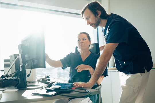 Female doctor discussing medical record with male coworker on computer while sitting in office at hospital