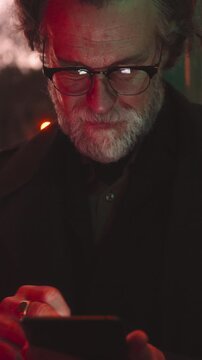 Senior man with glasses using smartphone app on city street at dusk. Reflections on his glasses suggest focus, possibly navigating taxi app or managing online banking in a busy urban environment
