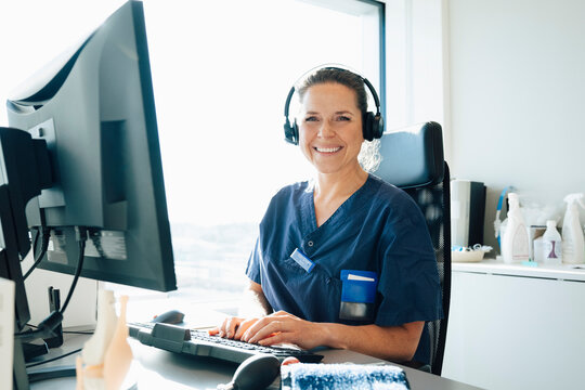 Portrait of smiling female doctor wearing headset with hand on keyboard sitting at desk in hospital office