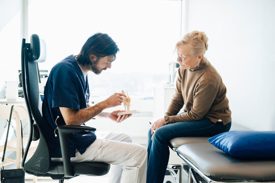 Male medical expert explaining joint condition to senior patient through anatomical model in hospital room