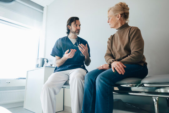 Low angle view of male medical professional discussing with senior woman while sitting on table in examination room at h